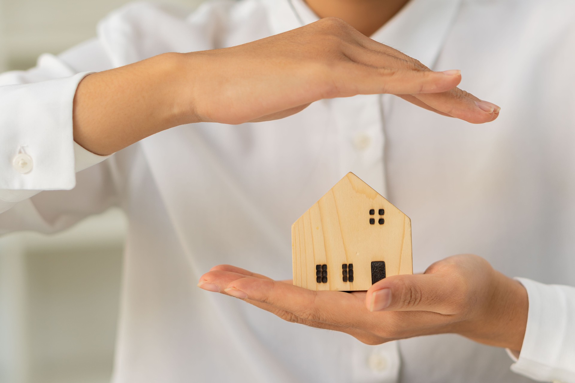 Die Hand einer asiatischen Frau hält ein kleines Hausmodell und verschränkt ihre Arme zur Verteidigung über dem Versicherungskonzept. Immobilien- und Pflegeheimpflege- und Familienkonzepte.